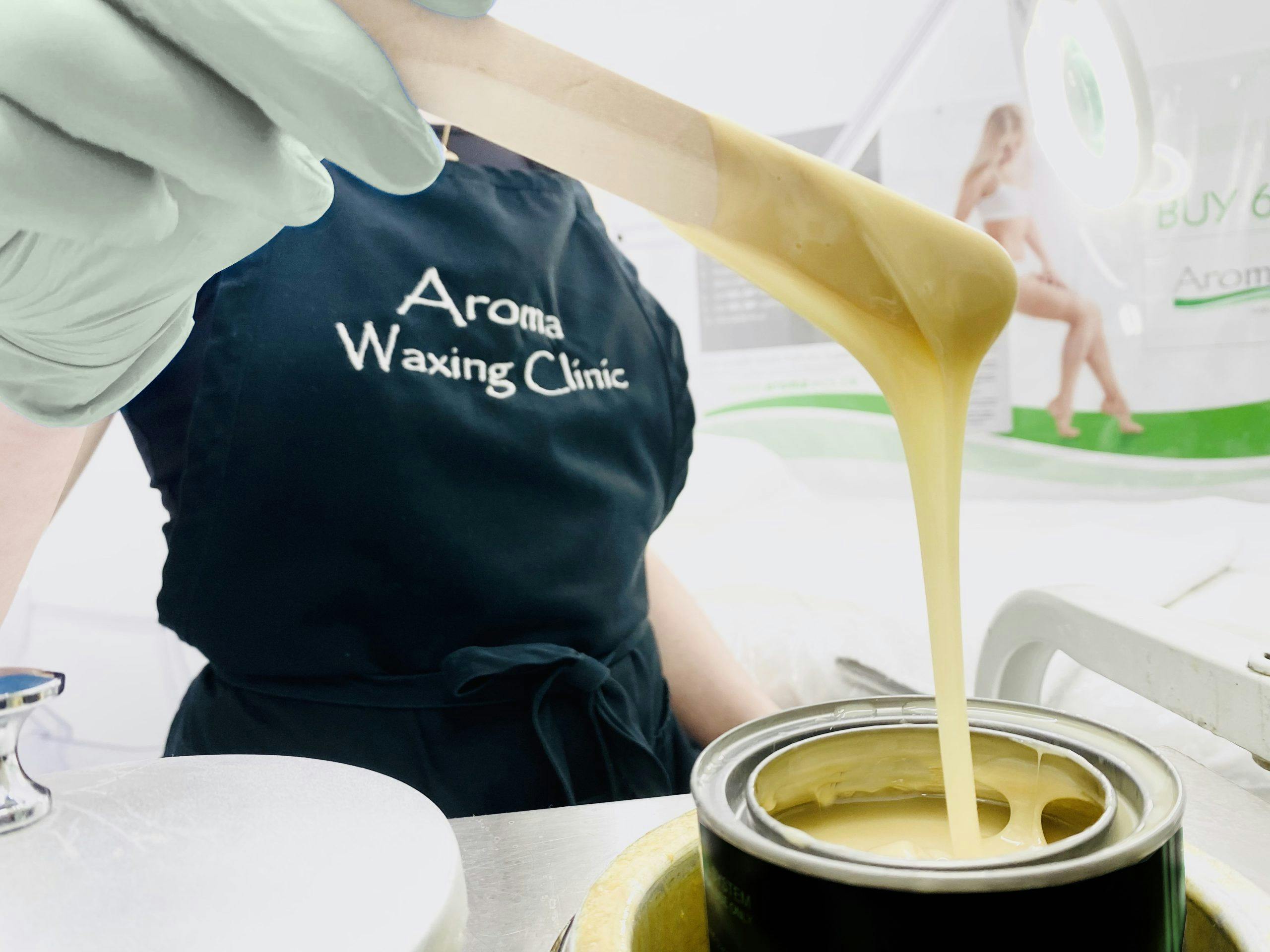 Person with Aroma Waxing Clinic apron pulling cream based wax with stick from wax pot