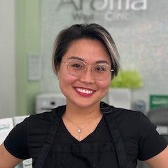 With a welcoming grin, Hong positions herself in her professional gear against the backdrop of the Aroma Waxing Clinic's insignia, symbolizing her fun-loving and comprehensive beauty philosophy